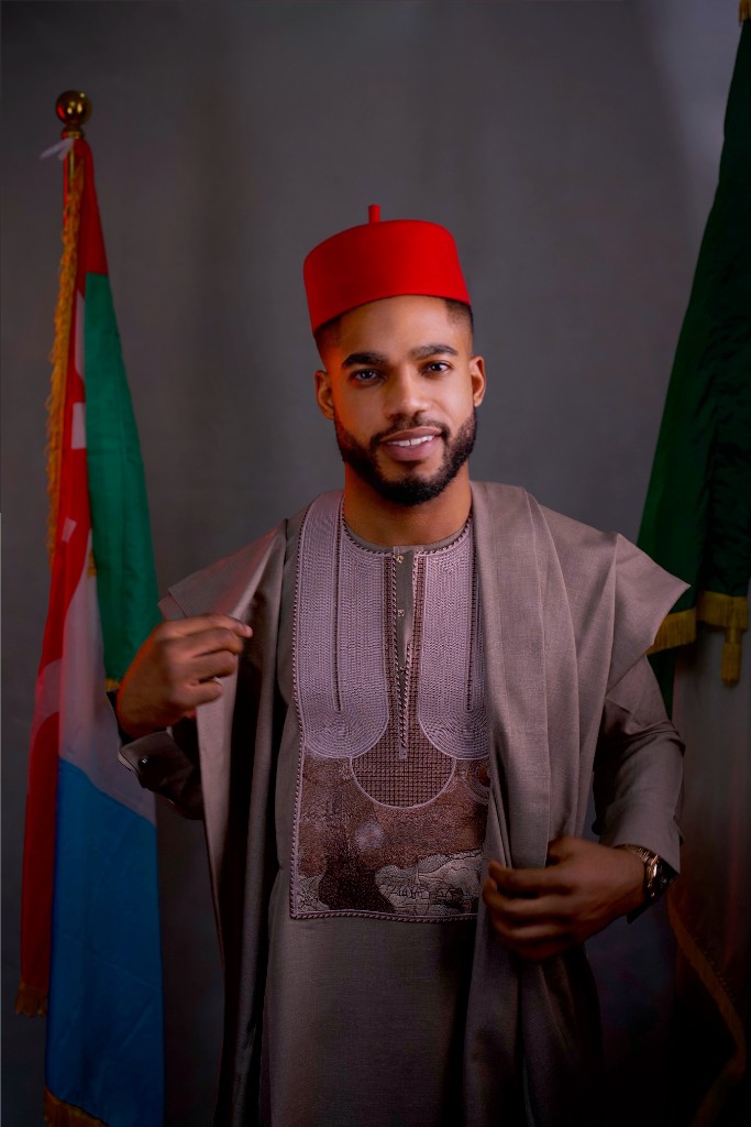 Osborn Nweze Umahi standing in a taupe agbada and red cap, holding the cloth of the robe.